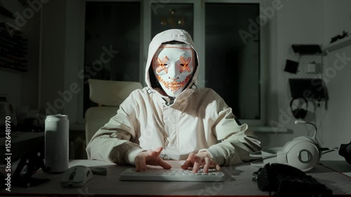 Close-up of masked hacker in hood typing at desk with headset and energy drink, illuminated in darkness. Reflects themes of cybersecurity, hidden identity and cyber threats.
