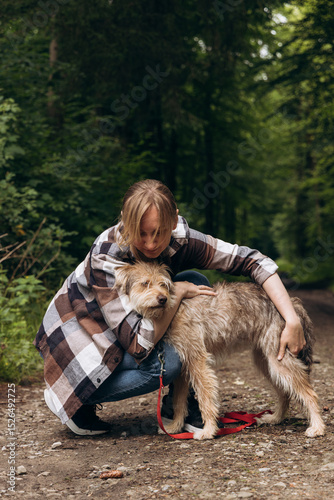 Woman in plaid shirt kneeling on forest path, lovingly embracing her dog, surrounded by lush greenery and natural beauty in a serene outdoor setting