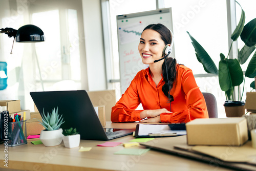 Smiling female entrepreneur working on laptop at her desk with packages in a bright modern office environment photo