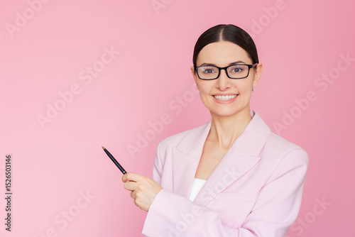 Young pretty Caucasian businesswoman in a pink suit and glasses pointing with pen at an empty pink background, looking at camera and smiling. Copy space