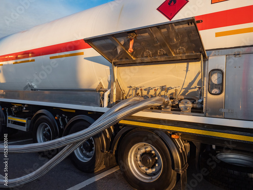 Fuel tanker truck with hazardous material sign, connected stainless steel hoses, control valves, and pressure gauges for liquid transfer – Logistics, Industrial Transport.