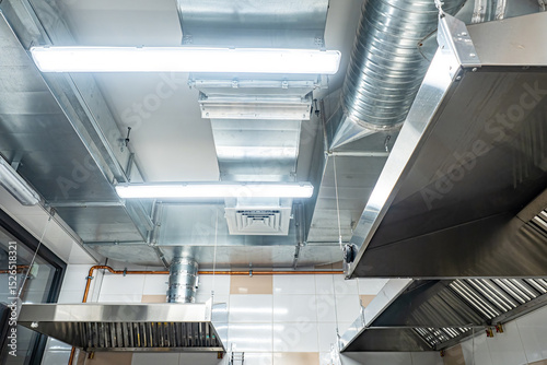 Industrial kitchen ceiling with stainless steel ventilation hoods, air ducts, and bright fluorescent lighting, ensuring proper air circulation. – Catering. Restaurant. Food Industry.