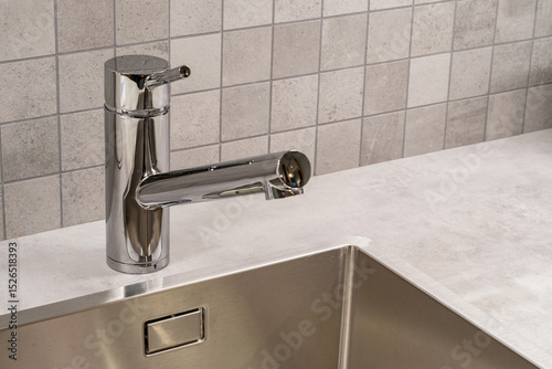 Close-up of a modern chrome kitchen faucet and stainless steel sink on a light countertop with tiled backsplash, minimal and clean design. – Interior Design. Home. Hospitality.