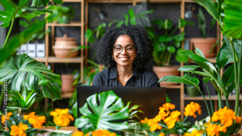 Smiling woman with curly hair working on laptop surrounded by vibrant plants and flowers