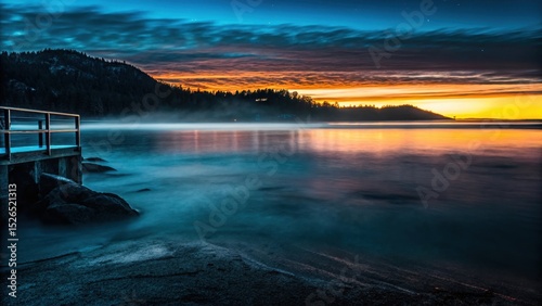 Serene Sunrise Pier: A tranquil lakeside scene at sunrise, showcasing a wooden pier extending into calm, mist-shrouded water, with a picturesque mountain range silhouetted against a vibrant.