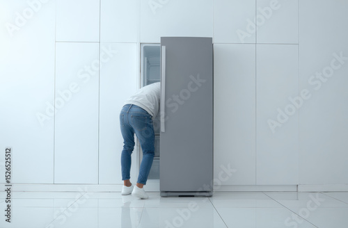 In a bright, modern kitchen, a man in jeans and white socks leans into a refrigerator, searching for something cool to beat the extreme summer heatwave