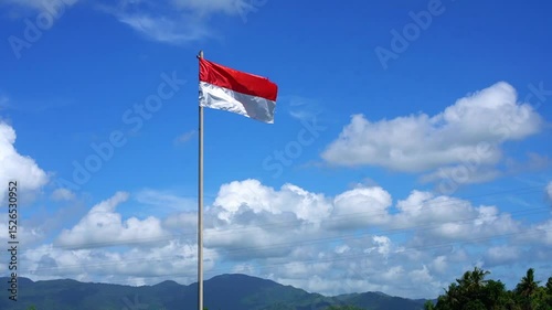 The red and white flag fluttering in the wind on a sunny day. Indonesian flag with blue sky, white clouds and mountains in the background.