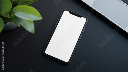 Modern smartphone with blank white screen lying on sleek black desk next to a silver laptop and green plant for technology, communication, and business themes
