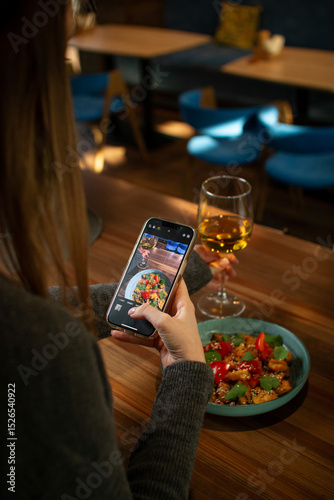 A young woman photographs her food with a smartphone while dining at a restaurant. A glass of white wine and a vibrant dish with vegetables are on the table