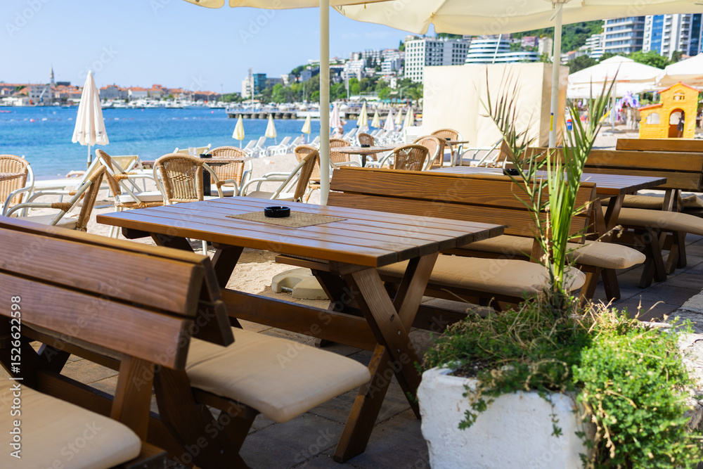 Obraz premium Wooden benches and tables at beach cafe under umbrella. Coastal hospitality, summer relaxation, and outdoor seating near sea.