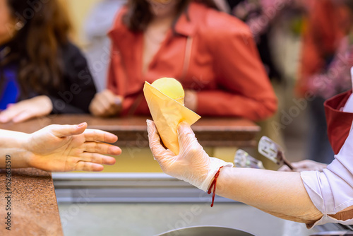 Ice cream seller giving ice cream cone to customer. Vendor in glove handing yellow ice cream cone to customer across counter. Concept of ice cream, food service, organic summer refreshment