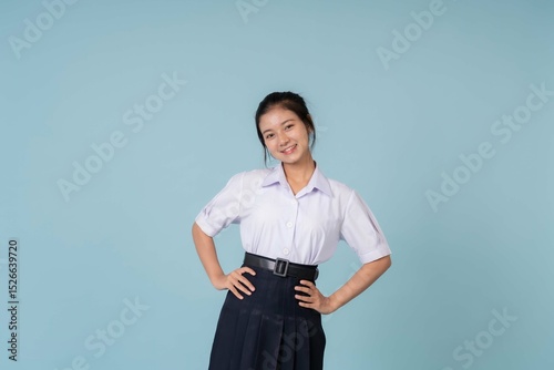 With a soft smile, the Asian school girl is posing in her cute high school uniform, her pretty face framed by the serene blue studio background that highlights her gentle personality.