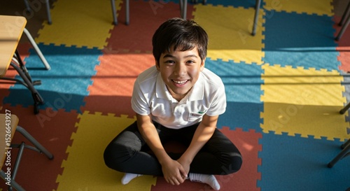 Young boy smiles while seated on a colorful school floor with desk legs surrounding him. Learning time.