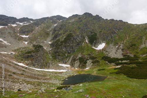 Mountain lake nestled in alpine valley with snow patches on rocky peaks and green meadows under cloudy sky