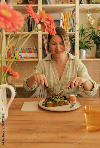 Crop middle-aged woman wearing stylish outfit eating delicious breakfast including egg and bacon with freshly greens served on wooden table in sunny outdoor cafe. Part of a series