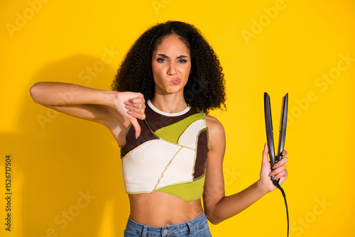 Young woman with curly hair expressing dislike for hair straightener tool on a vibrant yellow background