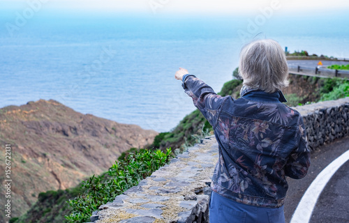 Back view of senior gray haired traveler woman in outdoor excursion looking at landscape and ocean view from top of hill. Horizon over water