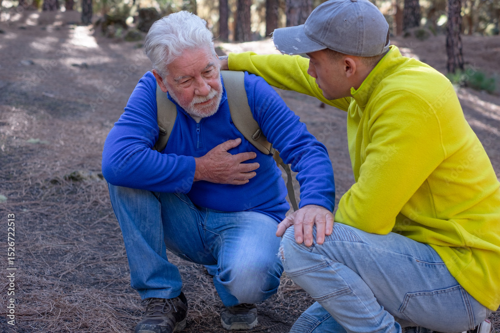 Fototapeta premium Old senior man hiking in the mountains with his young nephew pauses out of breath touching his heart. Elderly grandfather feeling unwell and his grandson assists him