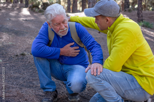 Old senior man hiking in the mountains with his young nephew pauses out of breath touching his heart. Elderly grandfather feeling unwell and his grandson assists him