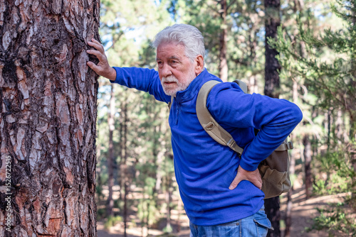 An elderly man out of breath during a mountain hike leaned against a tree trunk to recover. Active grandfather with beard and healthy lifestyle of a retired man