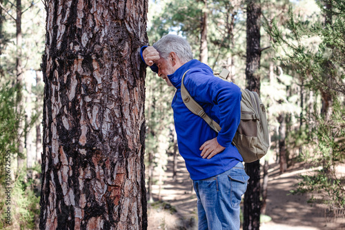 An elderly man out of breath during a mountain hike leaned against a tree trunk to recover. Active grandfather with beard and healthy lifestyle of a retired man