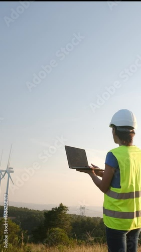 Female engineer in safety gear uses a laptop to monitor wind turbine performance in a field. Renewable energy and technology integration showcased in an outdoor setting