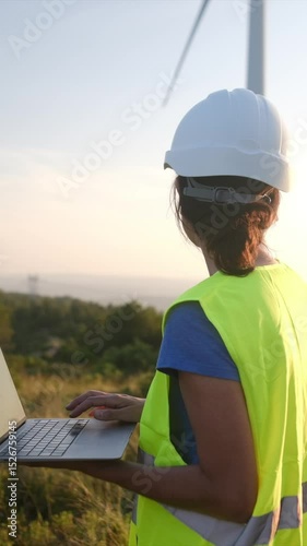 Female engineer in safety gear uses a laptop to monitor wind turbine performance in a field. Renewable energy and technology integration showcased in an outdoor setting