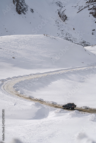 A black vehicle drives on a winding road covered in snow, surrounded by vast, pristine snow capped mountains under a cloudy sky