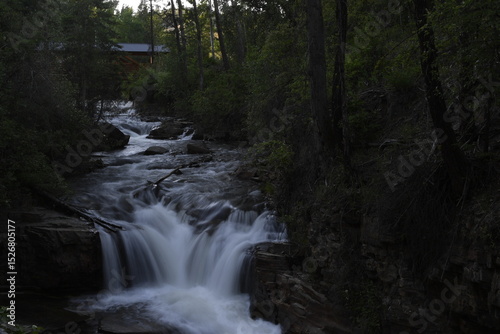 Slow shutter speed flowing creek water over rocks giving silken look with green foliage on sides and wooden bridge in background