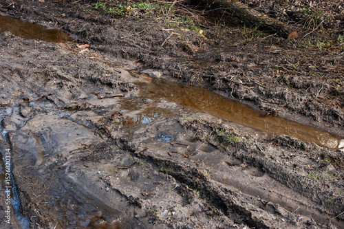 Very muddy dirt road with deep puddles and flowing water. The surface is churned up and full of footprints and tire tracks. The water is orange or brown from the clay soil.