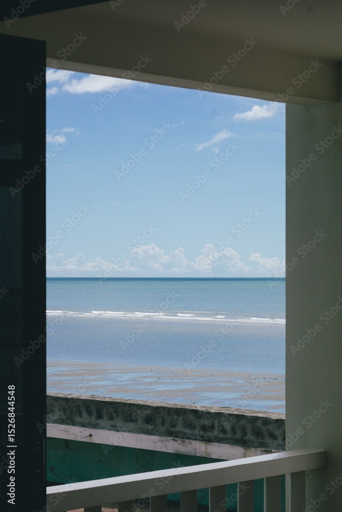 Fototapeta premium a view of the sea framed by an open doorway or window from what appears to be a room or balcony. The deep blue ocean stretches to the horizon under a bright sky with scattered white clouds.