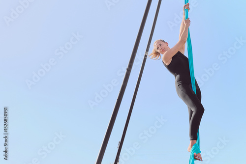 Young sporty blonde woman wearing a black jumpsuit performs aerial silk dance suspended while standing, gripping the fabric hanging from a metal structure outdoors.