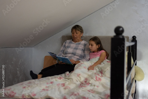 Grandmother reads a bedtime story to granddaughter in a cozy attic bedroom. The girl is tucked in bed, listening attentively to the story.