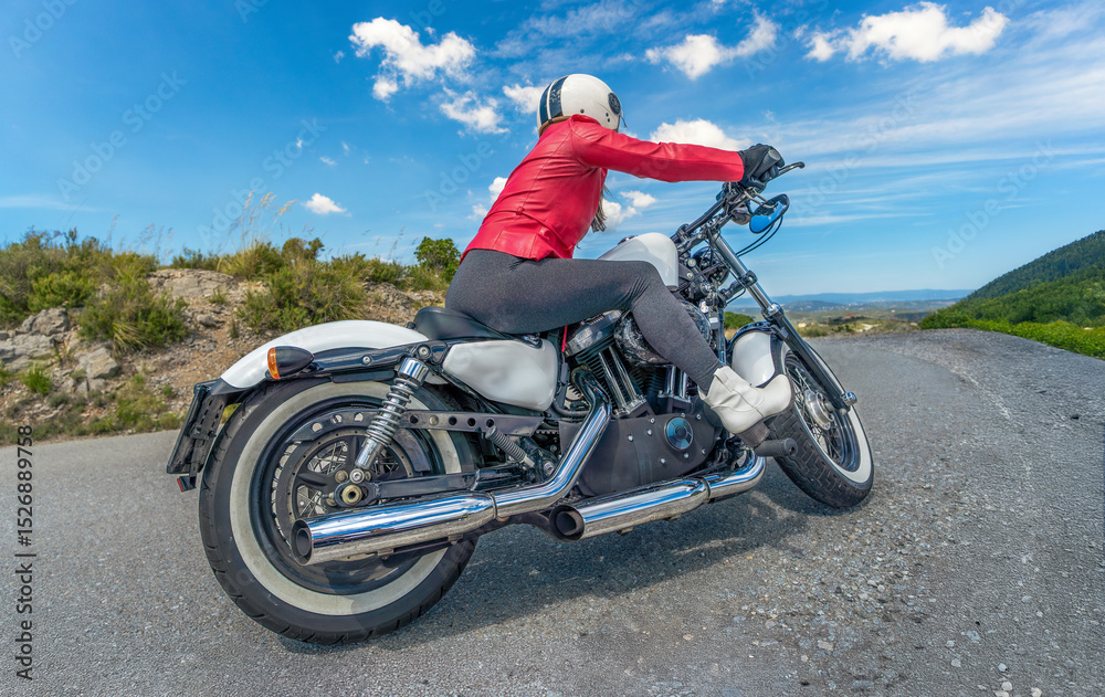 Obraz premium Side view of a female motorcyclist wearing a red jacket, tights, and white boots, riding her customized Harley-Davidson chopper motorcycle taking a hairpin turn along a scenic mountain road.
