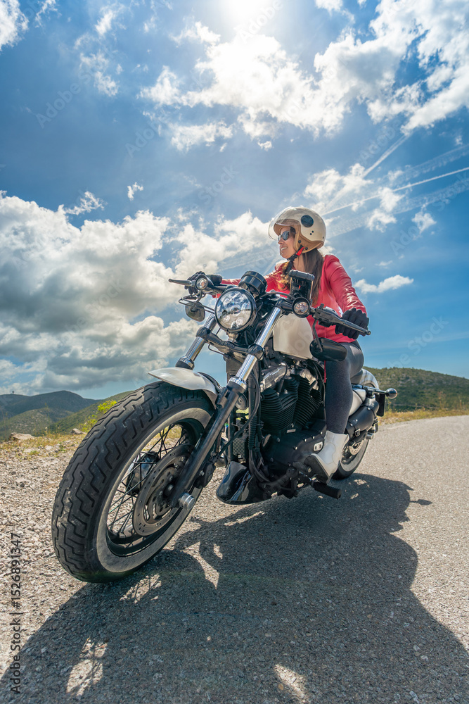 Fototapeta premium A young woman enjoying riding a classic chopper motorcycle looking at the mountains on a mountain road under a sunny sky with clouds illuminated by the sun.