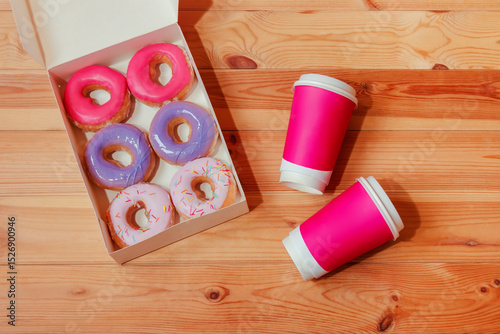 Pink paper coffee cups and donuts in paper box on wooden background.