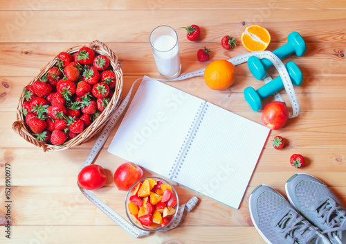 Empty notebook, yogurt, healthy fruit salad, wicker basket of fresh strawberry, dumbbells, sneakers and measuring tape on wooden surface.