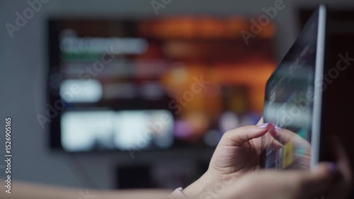 Relaxed woman browsing streaming service on tablet, reclining on sofa with blurred television screen behind, depicting casual home entertainment moment
