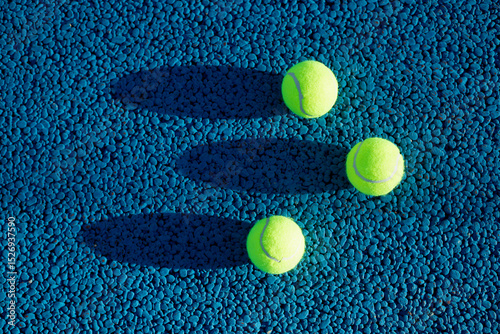 Billede på lærred Overhead view of three neon yellow tennis balls casting elongated shadows on a vibrant blue pebble-textured court surface, highlighting strong contrast and geometric composition