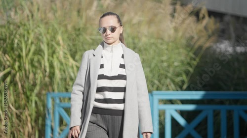 A self-assured woman walks confidently through a modern landscaped park. Her determined look and posture symbolize empowerment, high self-esteem, and main character energy.