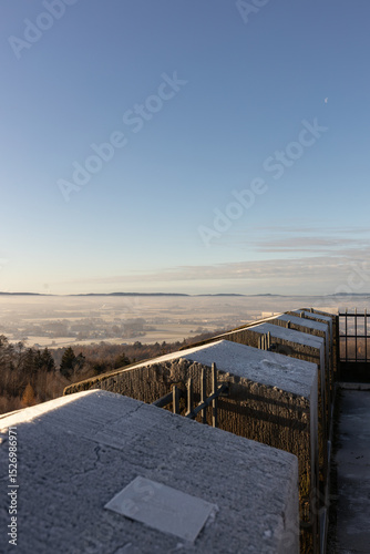 Frost-covered stone railing overlooking a misty valley, with a clear blue sky and distant mountains, creating a serene winter landscape that invites contemplation and appreciation of nature's beauty