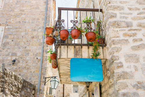 Fototapeta Naklejka Na Ścianę i Meble -  Small balcony with flower pots and blank blue sign on stone building copy space. Mediterranean charm, outdoor decor and vintage architecture.