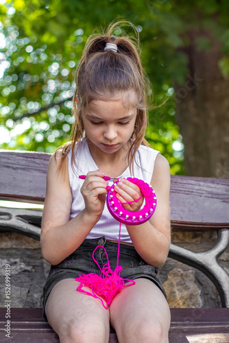 Young girl knitting with pink yarn on a park bench in summer
