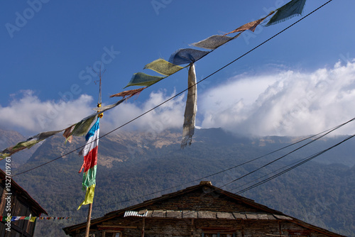 Brightly colored prayer flags sway in the breeze above rustic wooden buildings, with Annapurna mountains looming under a brilliant blue sky, creating a serene environment for trekkers photo
