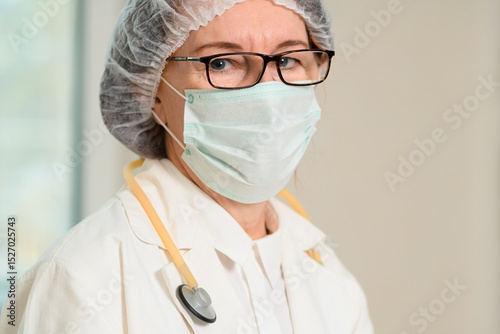 An experienced woman doctor in a white uniform and face mask stands in a hospital room. Her calm gaze and medical tools reflect care, readiness, and responsibility in healthcare service.