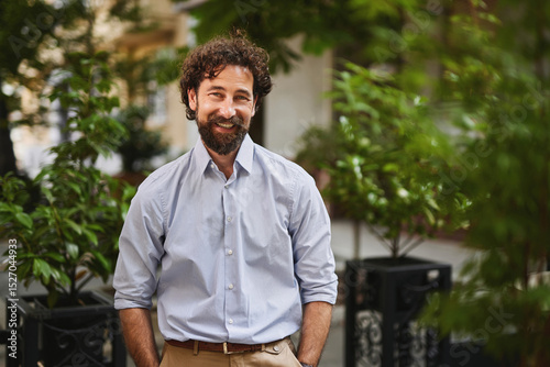 A man with curly hair and a beard stands in a vibrant outdoor area filled with plants, dressed in a light blue shirt. He looks relaxed and ready for business. photo