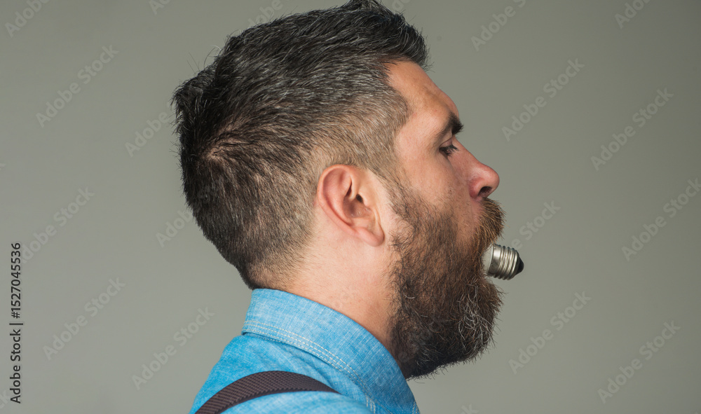 Obraz premium Bearded man with light bulb in mouth. Handsome man with stylish hairstyle in denim shirt eat light bulb lamp. Energy and electricity. Side view bearded man with electrical lightbulb in his mouth.