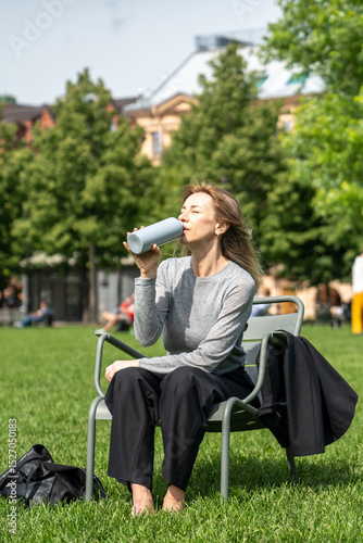 Exhausted woman squinting from heat, drinking water thirstily resting on grass in park after work. Refreshment, female on chair with beverage, park relief, hydration and recovery, sweltering weather