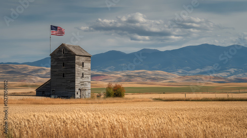 A rustic wooden grain elevator stands in a golden wheat field under a blue sky. An American flag waves proudly atop the structure, symbolizing Independence Day.