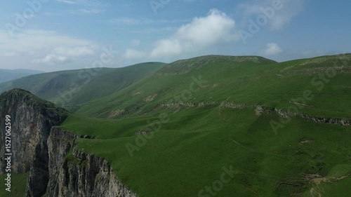 Beautiful panoramic view of green mountains with rocky formations, covered in lush grass, captured by drone flying backward against a clear blue sky.
